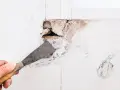 Mano con spatola rimuove intonaco da un buco nel muro troppo grande, rivelando mattoni e detriti.