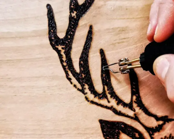 Dettaglio di un disegno a pirografia su legno: una mano guida le punte pirografo per creare le corna di un cervo.