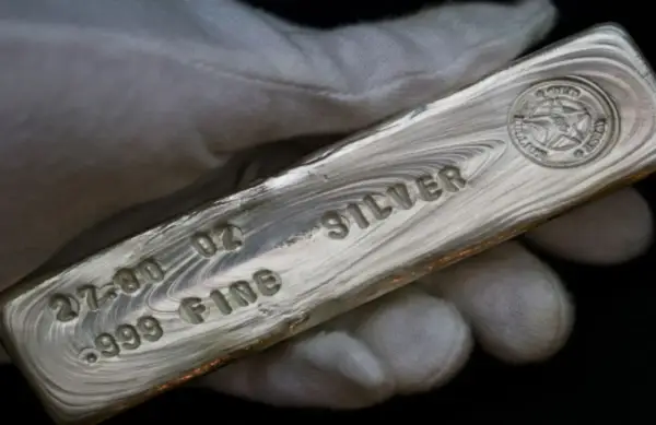 Mano guantata tiene un lingotto di **titolo argento** 999.9, con incisioni che indicano 27.00 oz e "FINE SILVER".