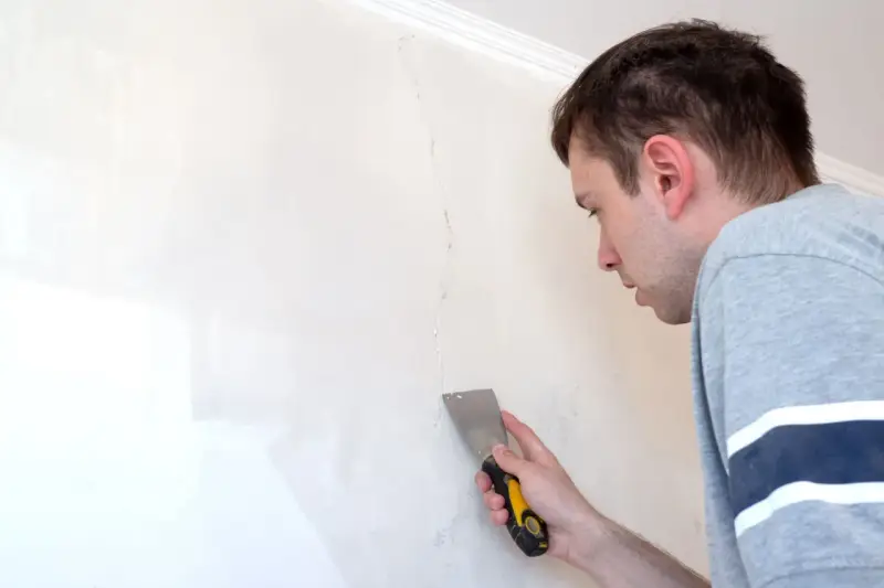 Uomo con spatola ripara una crepa su un muro bianco.