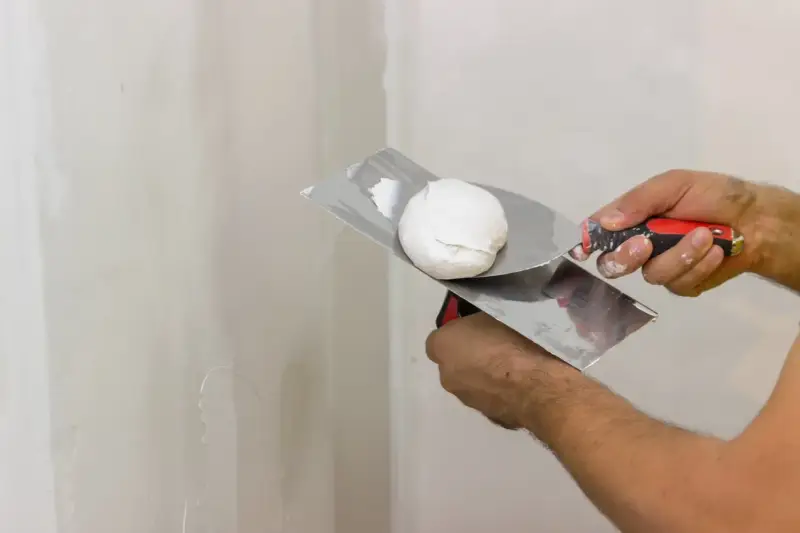 Mani che usano spatole per applicare stucco su un muro. Lo stucco serve a livellare e riparare le imperfezioni.