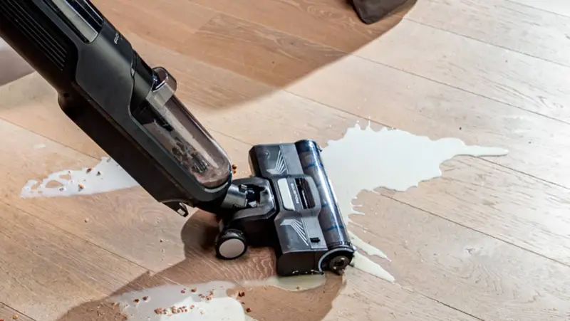 A wet/dry vacuum cleaner is cleaning up a spill of white liquid and debris on a wooden floor.