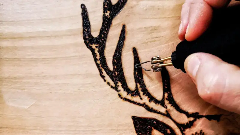 Dettaglio di un disegno a pirografia su legno: una mano guida le punte pirografo per creare le corna di un cervo.